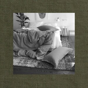 May include: A white bed with a gray linen duvet cover and a white pillow. A gray throw pillow is on the bed. The bed is in a bedroom with a white rug and a small table with a lamp.