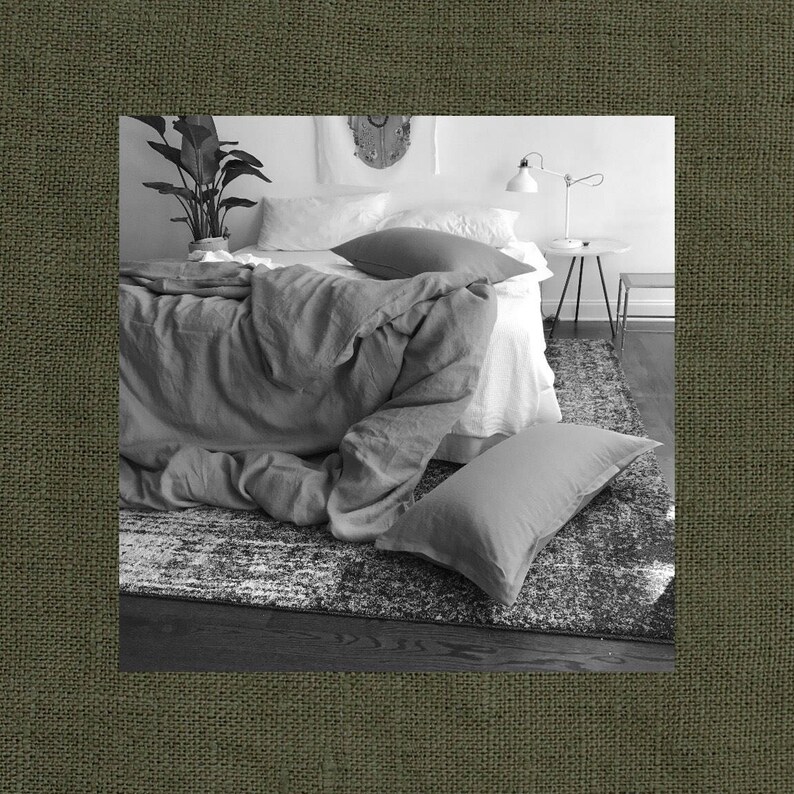 May include: A white bed with a gray linen duvet cover and a white pillow. A gray throw pillow is on the bed. The bed is in a bedroom with a white rug and a small table with a lamp.