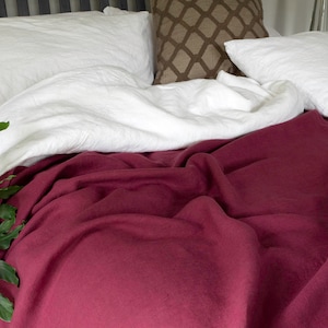 May include: A white linen duvet cover and pillowcases are folded on a bed with a deep burgundy linen throw blanket.