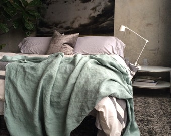 May include: A sage green linen throw blanket draped over a bed with white and gray striped bedding, a textured gray throw pillow, and a white adjustable desk lamp on a round side table, set against a wall with a large dark cloud art print.