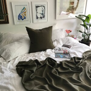 May include: A white bed with a green linen throw blanket and white linen pillows. There is a green pillow on top of the white pillows. A magazine is on the bed and a plant is in the background.