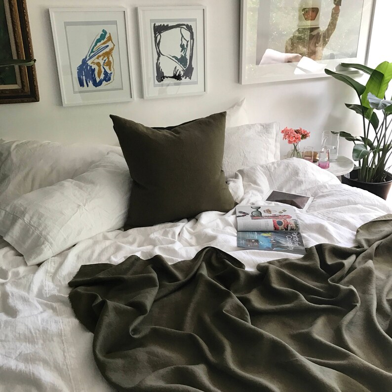 May include: A white bed with a green linen throw blanket and white linen pillows. There is a green pillow on top of the white pillows. A magazine is on the bed and a plant is in the background.