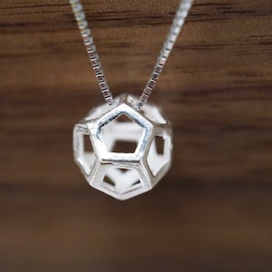Może przedstawiać: Srebrny naszyjnik z geometryczną zawieszką. Zawieszka ma pusty, trójwymiarowy kształt z pięciokątnymi otworami. Łańcuszek naszyjnika ma delikatny, kwadratowy splot. Tło stanowi rozmyta, drewniana powierzchnia.