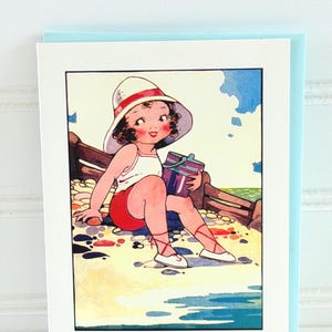 May include: A vintage-style greeting card featuring a cartoon illustration of a child at the beach. The child wears a white hat with a red band, a white top, and red shorts. The child is holding a striped box, with the ocean and sky in the background.
