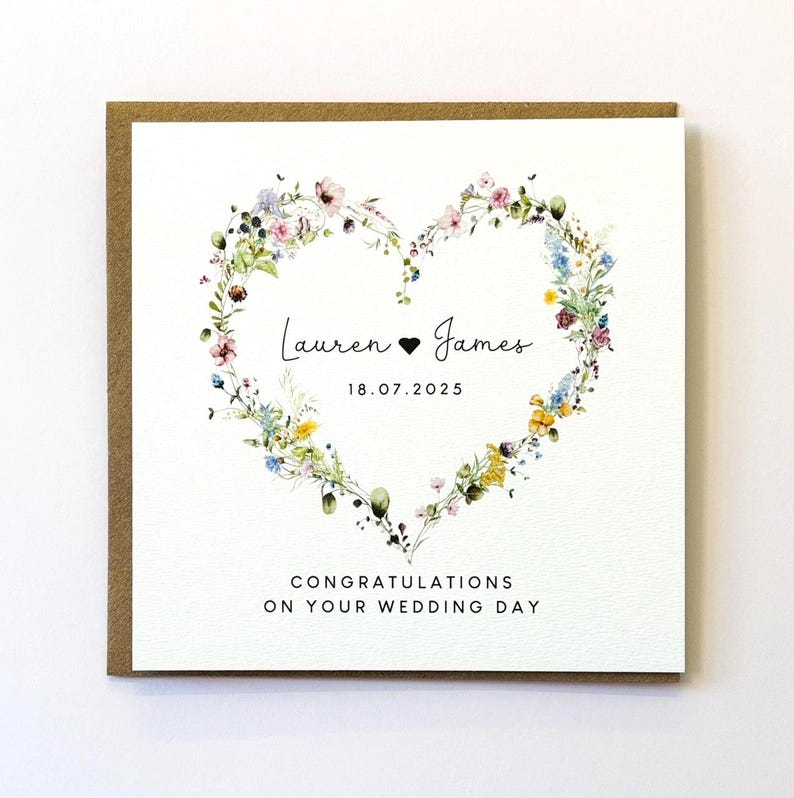 Pu&ograve; includere: Un biglietto di nozze bianco con un design a forma di cuore floreale. Il biglietto dice "Lauren &hearts; James 18.07.2025" e "Congratulazioni per il vostro matrimonio".
