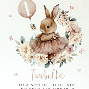 May include: A watercolour illustration of a bunny wearing a tutu and holding a balloon with the number 1. The bunny is surrounded by a floral wreath and butterflies. The text reads "Isabella, To a special little girl on your 1st birthday."