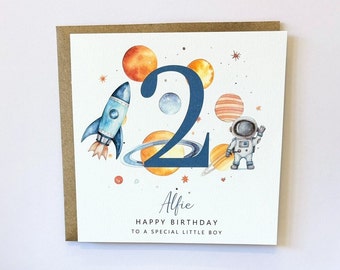 May include: A white card with a blue number 2 surrounded by planets and stars. An astronaut and a rocket ship are also on the card. The text "Alfie Happy Birthday to a special little boy" is printed on the card.
