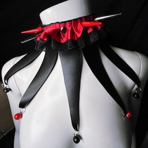 May include: A black choker with red satin ribbon and black lace trim. The choker has five black ribbon tails with red beads and silver bells at the ends. The choker also has silver spikes and red accents.