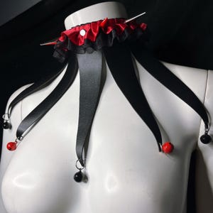 May include: A black choker with red and black ribbon accents and dangling black and red beads. The choker has a silver pin in the center.