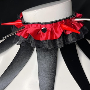 May include: A red and black choker with silver spikes and a ruffled trim. The choker is worn on a white mannequin.