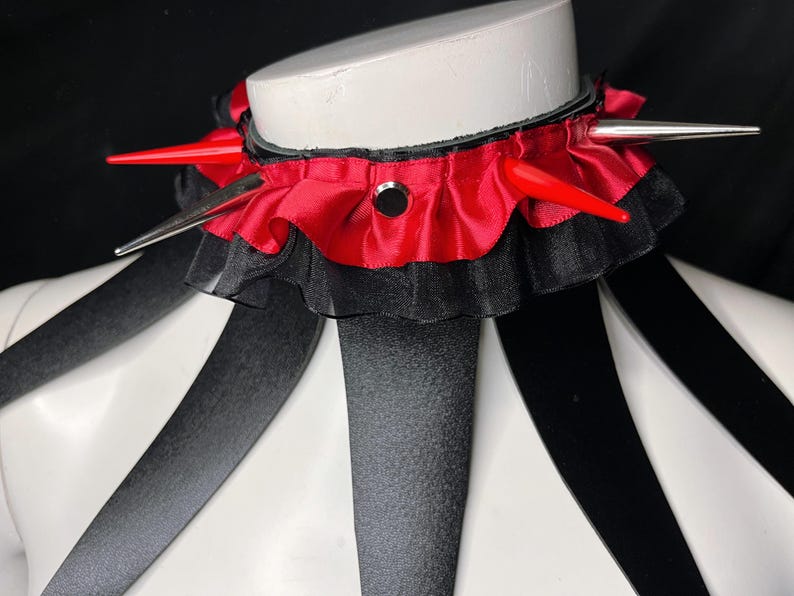 May include: A red and black choker with silver spikes and a ruffled trim. The choker is worn on a white mannequin.