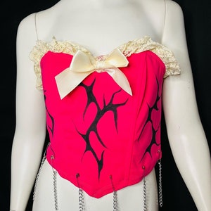 May include: A pink strapless corset with black tribal-style designs and a white satin bow. The corset has silver safety pins with chains attached to the bottom.