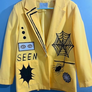 May include: A bright yellow blazer with black graphic designs. The blazer features a spiderweb, spiders, and the word "SEEN" in black lettering. Additional designs include three black dots and an eye graphic.