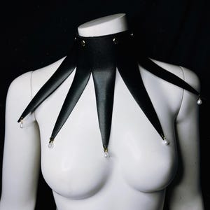 May include: A black leather choker with a starburst design and gold accents. The choker has five points that extend down the chest and are adorned with white pearl-like beads.