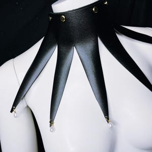 May include: A black leather choker with gold studs and pearl accents. The choker has five points that extend down from the collar.