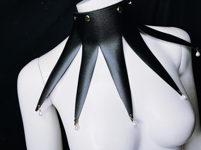 May include: A black leather choker with gold studs and pearl accents. The choker has five points that extend down from the collar.