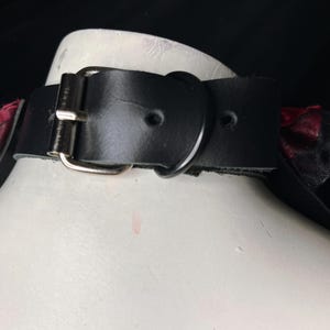 May include: Black leather choker with a silver buckle and two red and black bows on either side.