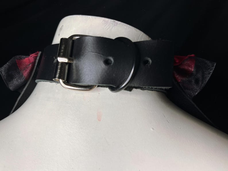 May include: Black leather choker with a silver buckle and two red and black bows on either side.
