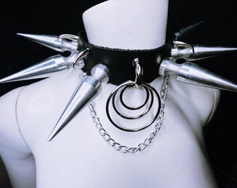 Könnte beinhalten: Ein schwarzer Leder-Choker mit silbernen Spikes und einer Kette mit mehreren silbernen Ringen.