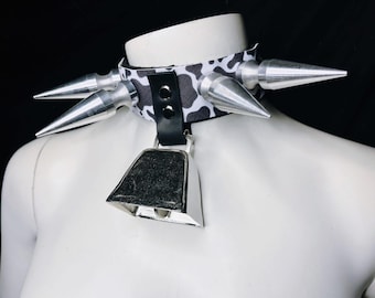 May include: A black and white cow print collar with silver spikes and a silver bell. The collar is made of fabric and leather, and is designed to be worn around the neck.
