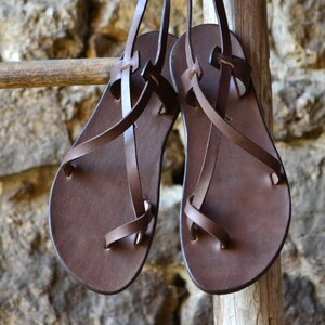May include: A pair of brown leather sandals with criss-cross straps. The sandals are hanging on a wooden ladder.