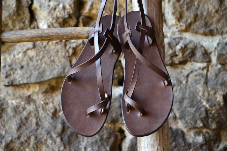 May include: A pair of brown leather sandals with criss-cross straps. The sandals are hanging on a wooden ladder.