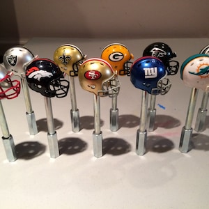 May include: A collection of miniature football helmets, each representing a different NFL team, are displayed on metal rods. The helmets are painted in team colors and feature team logos. The helmets include the New England Patriots, Denver Broncos, New Orleans Saints, San Francisco 49ers, Green Bay Packers, New York Giants, Philadelphia Eagles, Miami Dolphins, Dallas Cowboys, and the Oakland Raiders.