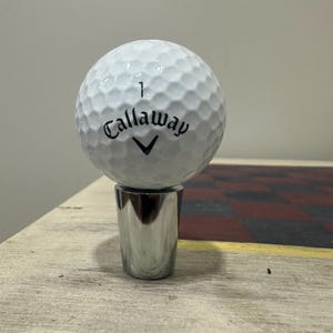 May include: A white Callaway golf ball with a dimpled surface and the number 1 printed on it, resting on a polished silver-colored metal stand. The golf ball features the black Callaway brand name and logo printed on the front side.