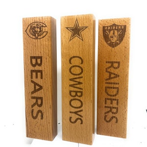 Könnte beinhalten: Drei Holzblöcke mit Teamlogos und Namen. Die Blöcke sind mit dem Logo der Chicago Bears und "BEARS", dem Stern der Dallas Cowboys und "COWBOYS" sowie dem Logo der Las Vegas Raiders und "RAIDERS" graviert.