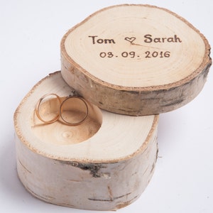 May include: A wooden ring box with a lid, made from a tree slice. The lid has the text "Tom ♥ Sarah 03.09.2016" engraved on it. Two gold rings are inside the box.
