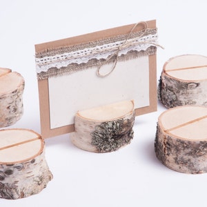 Puede incluir: Cinco soportes de número de mesa de madera rústica con un acabado natural de corteza de abedul. Cada soporte tiene una ranura para una tarjeta o un letrero. Los soportes están dispuestos sobre una superficie blanca.