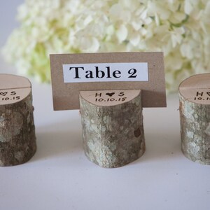 Puede incluir: Tres soportes de número de mesa de madera rústica con el texto "H♥S 10.10.15" grabado en cada uno. El soporte del medio tiene una tarjeta marrón con el texto "Mesa 2" en ella.