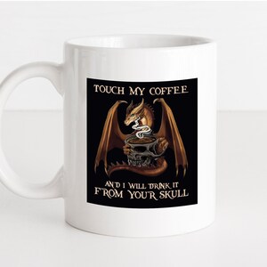May include: White ceramic coffee mug with a black and brown dragon illustration. The dragon is holding a coffee cup and has steam coming out of its nostrils. The text on the mug reads "Touch my coffee and I will drink it from your skull."