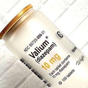 May include: A white cylindrical prescription bottle with a wooden-textured cap. The label reads "Valium (diazepam) 10 mg" and indicates the bottle contains 100 tablets. The bottle is set against a white brick background.