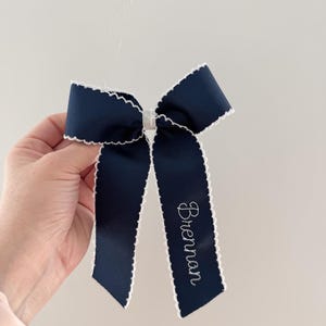 May include: Navy blue bow with white scalloped edges. The bow features the name "Brennan" embroidered in white script on one of the tails. The bow is held against a plain white background.