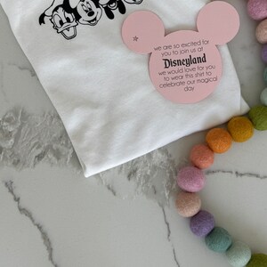 May include: White t-shirt with a black print of Mickey Mouse, Minnie Mouse, Donald Duck, Goofy, and Pluto. A pink card with the text "we are so excited for you to join us at Disneyland we would love for you to wear this shirt to celebrate our magical day" is on top of the shirt.