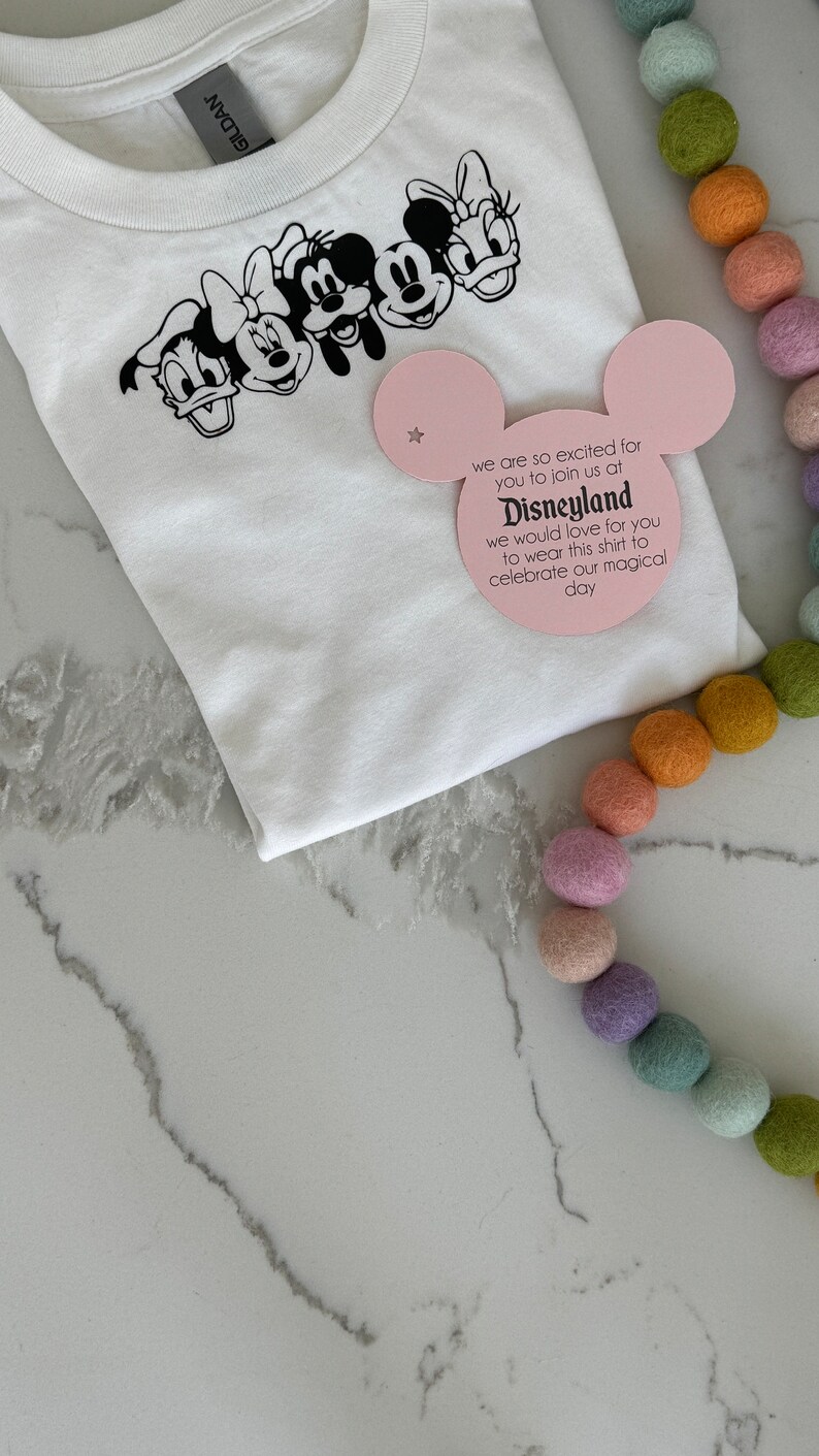 May include: White t-shirt with a black print of Mickey Mouse, Minnie Mouse, Donald Duck, Goofy, and Pluto. A pink card with the text "we are so excited for you to join us at Disneyland we would love for you to wear this shirt to celebrate our magical day" is on top of the shirt.