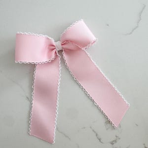 May include: A pale pink ribbon bow with white scalloped edging. The bow is tied in the centre with a white band and has two long, flowing tails. The ribbon has a ribbed texture.