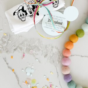 May include: A white t-shirt with a black and white image of Mickey Mouse, Minnie Mouse, and Donald Duck. The shirt is tied with a colorful ribbon and a white tag with the text "We are so excited for you to join us at Disneyland. We would love for you to wear this shirt to celebrate a magical day."