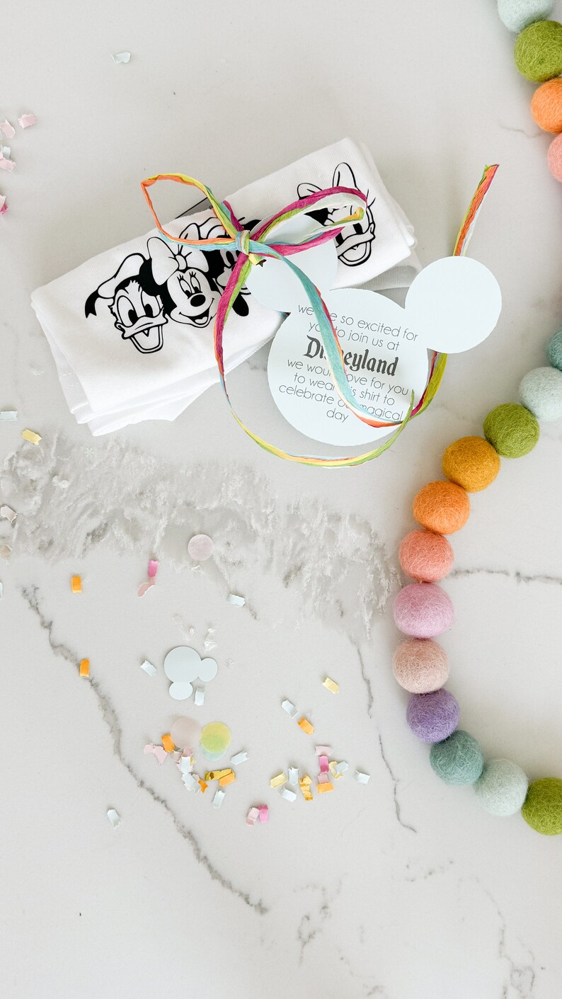 May include: A white t-shirt with a black and white image of Mickey Mouse, Minnie Mouse, and Donald Duck. The shirt is tied with a colorful ribbon and a white tag with the text "We are so excited for you to join us at Disneyland. We would love for you to wear this shirt to celebrate a magical day."
