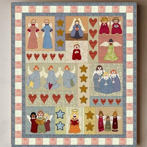 Könnte beinhalten: Ein farbenfroher Patchwork-Decken mit einem sich wiederholenden Muster aus Engeln und Herzen. Die Decke ist gerahmt und hängt an einer hellgrauen Wand über einem Holztisch und einem roten Stuhl.