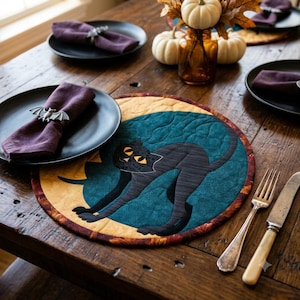 Op de afbeelding: Een ronde gewatteerde placemat met een silhouet van een zwarte kat tegen een gele maan en een groenblauwe achtergrond, geplaatst op een houten tafel met zwarte borden, paarse servetten, zilveren vleermuis servetringen, een vork, mes en witte pompoenen.