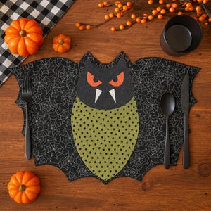 Può includere: Un'apparecchiatura da tavola a tema Halloween con un sottopiatto a forma di pipistrello con un motivo a ragnatela. Il pipistrello ha occhi arancioni e un corpo verde. Posate nere, zucche e una ciotola nera completano l'arrangiamento festivo.