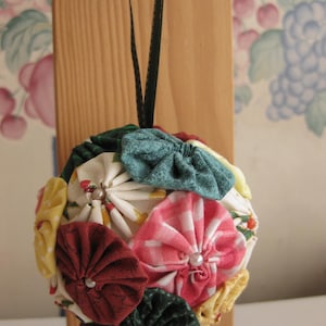 Op de afbeelding: Een handgemaakte kerstbal gemaakt van stofresten in verschillende kleuren en patronen, waaronder rood, groen, geel, wit en roze. De bal is rond en heeft een lus om op te hangen.