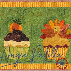 May include: A row of five decorated cupcakes with various fall-themed toppings, including a pilgrim hat, a pumpkin, a turkey, a carrot, and fall leaves. The cupcakes are on a green background with a yellow and orange striped border. The text "Angie Padilla" is written in cursive at the bottom of the image.