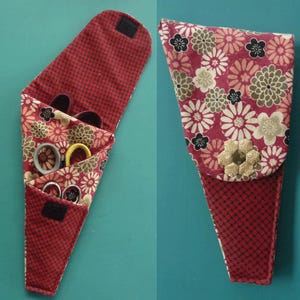 May include: A red and black fabric scissor holder with a floral pattern. The holder has a flap that closes with a velcro strap. The holder is filled with scissors and other tools.