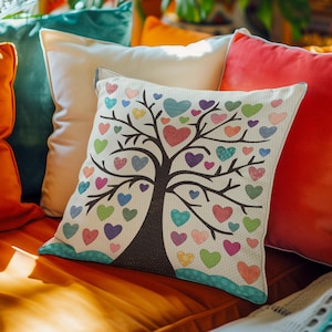 May include: A decorative cushion with a white background featuring a black tree with colourful hearts on its branches. The cushion is on a sofa with other cushions in various colours.