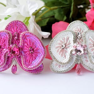 May include: Two orchid brooches, one pink and one white, are displayed. The pink brooch is covered in sequins and features a large pink crystal. The white brooch has pink accents and a teardrop crystal.