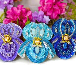 May include: Three beaded iris flower brooches in shades of purple, turquoise, and blue. Each brooch is embellished with sequins and crystals, and features a yellow center. The brooches are set against a backdrop of pink and purple flowers.