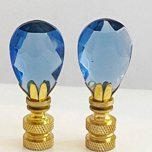 May include: Two blue glass finial lamp finials with gold metal bases. The finials are shaped like teardrops and have a faceted design.